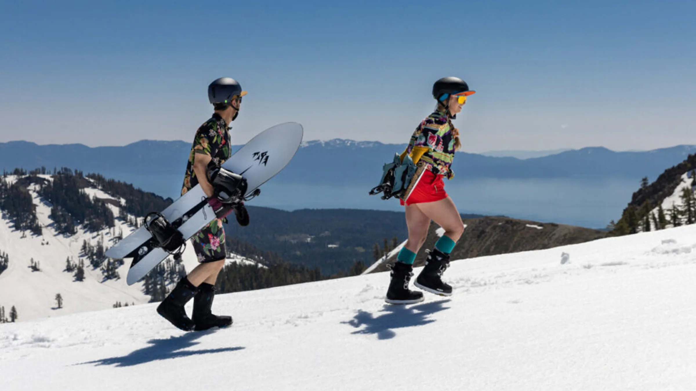 Two snowboarders in spring attire, hiking with their snowboards on a mountain ridge with Lake Tahoe in the background.
